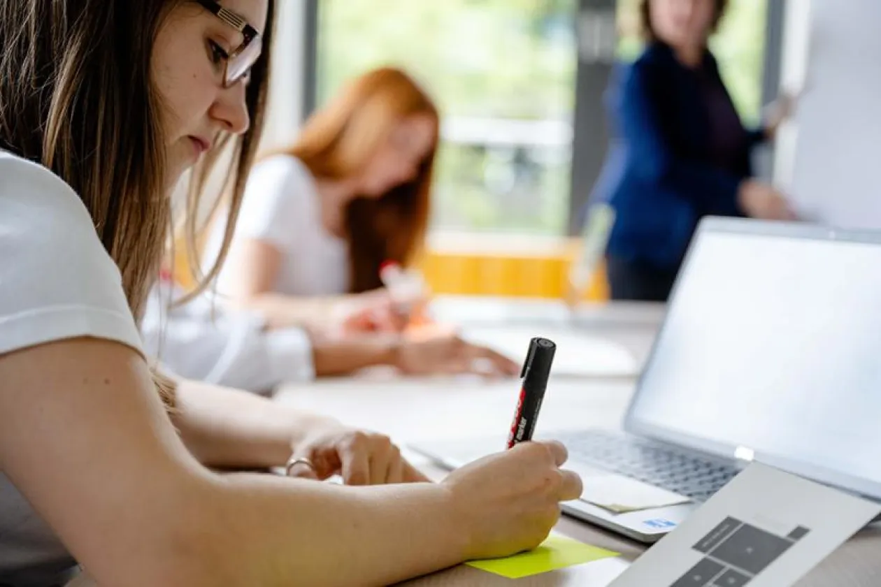 Studentin schreibt etwas. Im Hintergrund ist eine Lehrperson zu sehen. 
