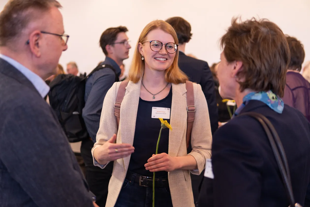 Foto: Drei Personen stehen im Kreis und unterhalten sich. Zwei Personen sind schräg von hinten zu sehen. Im Fokus steht eine lächelnde Frau mit einer gelben Blume in der Hand.