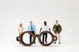 Eine Brille groß im Vordergrund mit diversen Menschen unterschiedlicher Haut- und Haarfarbe verschwommen und klein im Hintergrund.