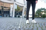 Eine Person tastet mit einem weißen Langstock das Pflaster vor dem Audimax ab – Symbol für Barrierefreiheit und Inklusion auf dem Campus.