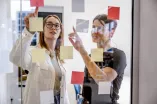 Fot: Zwei Frauen stehen hinter einer Glaswand und zeigen beide auf eines der vielen bunten Post-its, die an dieser befestigt sind.