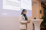 Foto: Eine Frau hält Moderationskarten in der hand und steht auf der Bühne. Sie spricht.