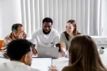 Foto: Mehrere Personen sitzen an einem Tisch mit Laptops und Stiften. Sie unterhalten sich, schauen auf die Laptops und scheinen zu überlegen.