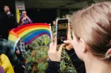 Foto: Eine Person mit einer Regenbogen-Fahne in der Hand macht ein Foto mit Smartphone von zwei Personen die erhöht/auf einer Bühne mit einem Mikrofon sprechen.