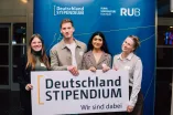 Foto: Vier Personen stehen vor einem blauen Aufsteller. Auf dem Aufsteller befinden sich das Logo des Deutschlandstipendiums und das Logo der Ruhr-Universität Bochum. Die vier Personen halten zusammen ein Schild mit der Aufschrift "Deutschlandstipendium - Wir sind dabei".
