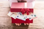 Foto: Zwei Hände halten eine rote Geschenkbox mit roter Schleife. Der Deckel der Box ist leicht geöffnet. Durch den Deckel sieht man Geldscheine.