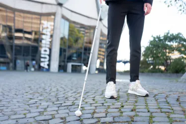 Eine Person tastet mit einem weißen Langstock das Pflaster vor dem Audimax ab 