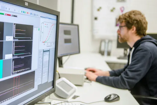 Das Bild zeigt im Hintergrund einen Mann am Schreibtisch am PC, im Vordergrud einen Bildschirm mit Graphen.