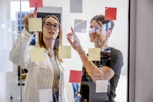 Fot: Zwei Frauen stehen hinter einer Glaswand und zeigen beide auf eines der vielen bunten Post-its, die an dieser befestigt sind.