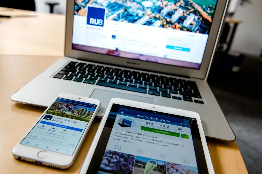 Ein Laptop, ein Handy und ein Tablet, jeweils eingeschaltet, liegen auf dem Schreibtisch