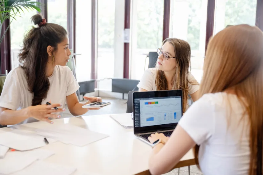 Foto: Drei Studentinnen sitzen an einem Tisch mit Papieren, Stiften, Taschenrechner, Laptop. Sie sprechen miteinander. Auf dem Laptop sind bunte Statistiken zu sehen.