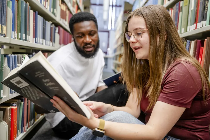 Foto: Zwei Studierende sitzen zwischen Bücherregalen auf dem Boden in der Bibliothek und lesen gemeinsam in einem Buch.