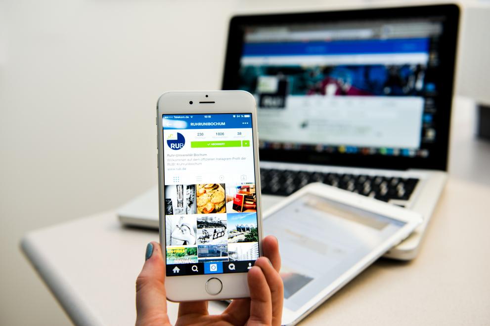 Eine Hand hält ein Handy, darauf zu sehen die RUB-Instagram-Seite, im Hintergrund Laptop und Tablet auf dem Tisch liegend.