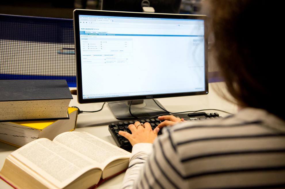 Eine Person sitzt in der Bibliothek am Computer und tippt, neben der Tastatur liegen aufgeschlagene Bücher.