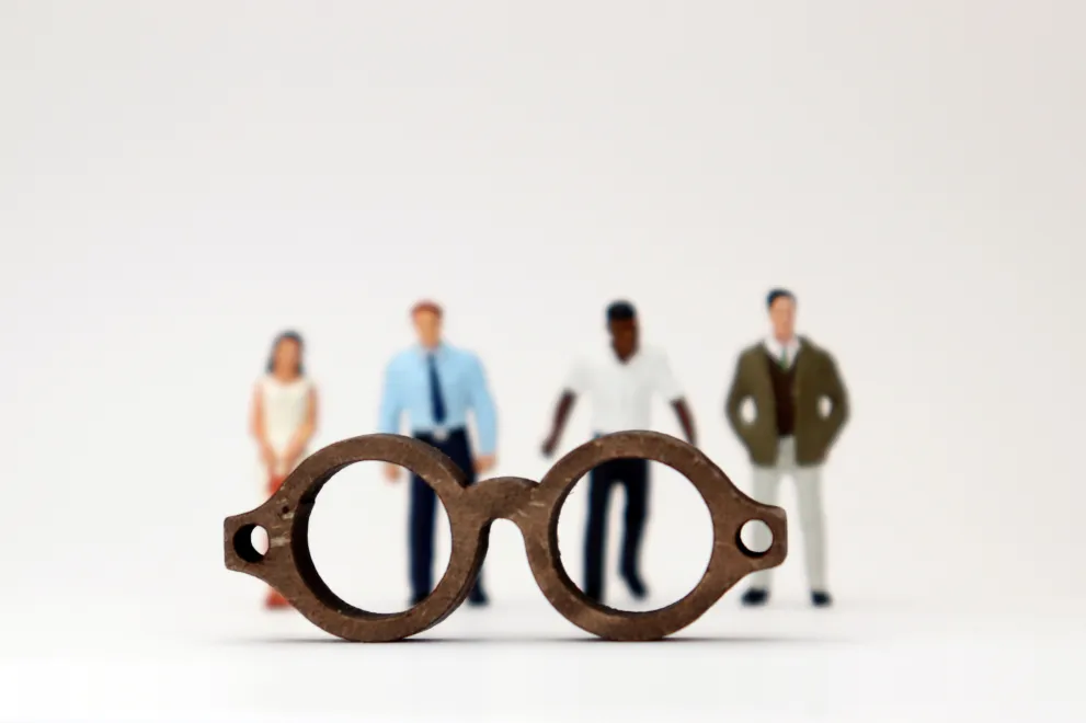 Eine Brille groß im Vordergrund mit diversen Menschen unterschiedlicher Haut- und Haarfarbe verschwommen und klein im Hintergrund.