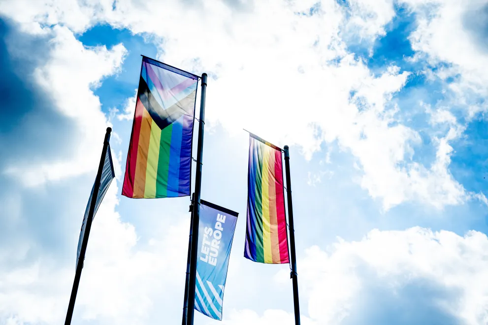 Das Bild zeigt zwei Regenbogenflaggen vor blauem Himmel, außerdem eine Flagge mit dem Logo von "Let´s Europe"