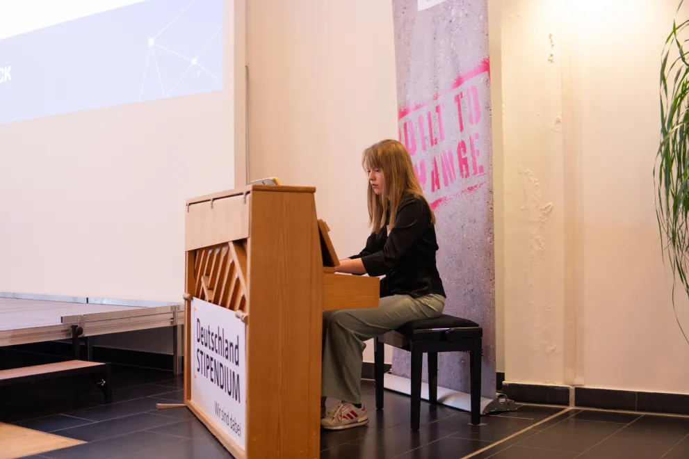 Foto: Eine Frau sitzt an einem Klavier und spielt. An der Front des Klaviers ist in Form eines großen Banners das Logo des Deutschlandstipendiums angebracht.