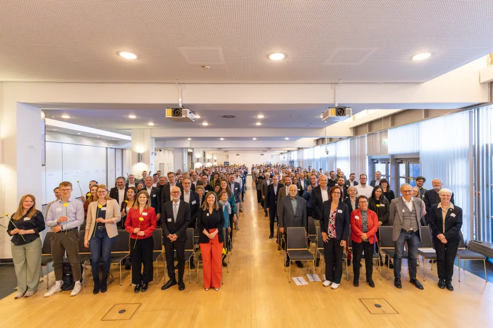 Foto: Gruppenfoto ins Plenum einer Veranstaltung. Alle Teilnehmenden stehen und lächeln in die Kamera.