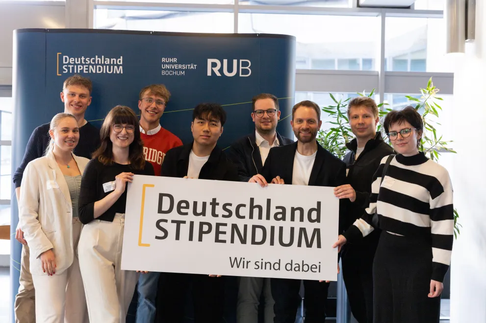 Foto: Einige Personen stehen und gucken lächelnd in die Kamera. Sie halten ein Banner mit dem Logo des Deutschlandsstipendiums auf Bauchhöhe.