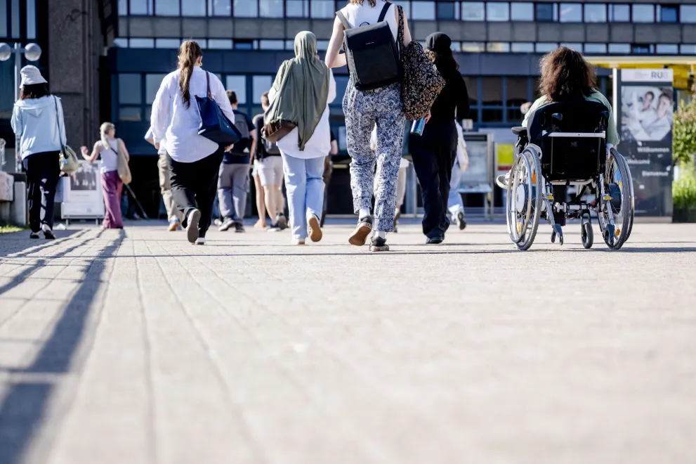 Verschiedene Menschen überqueren die Unibrücke, darunter eine Rollstuhlfahrerin, sie sind von hinten zu sehen