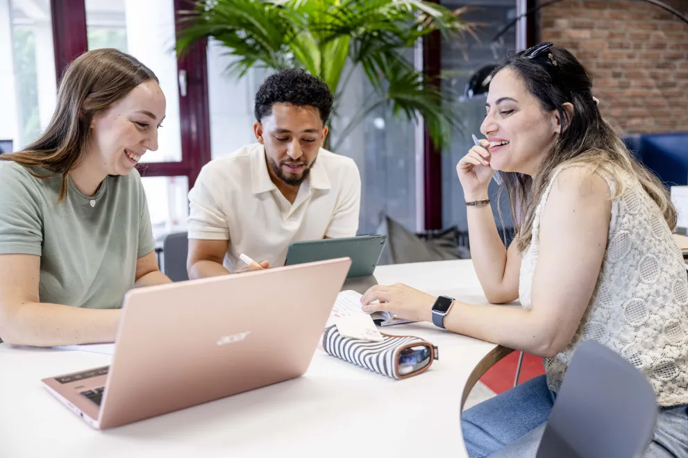drei Studierende sitzem am Tisch, unterhalten sich und schauen auf Laptops