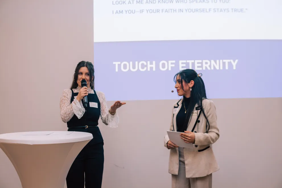 Foto: Zwei Frauen stehen nebeneinander. Eine Person hält ein Mikrofon, gestikuliert und spricht gerade. Die andere Person hält Moderationskarten in der Hand und guckt die sprechende Person an.