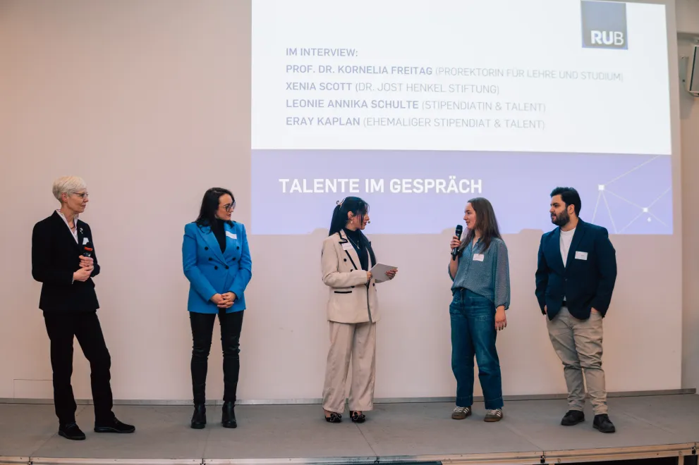 Foto: Fünf Personen stehen nebeneinander auf einer Bühne. Die Person in der Mitte hält Moderationskarten in der Hand.