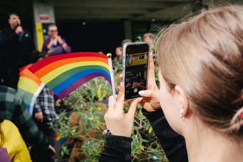 Foto: Eine Person mit einer Regenbogen-Fahne in der Hand macht ein Foto mit Smartphone von zwei Personen die erhöht/auf einer Bühne mit einem Mikrofon sprechen.