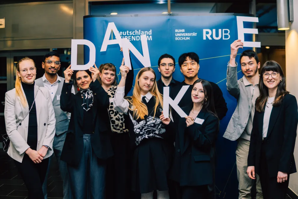 Foto: Mehrere Personen stehen vor einem blauen Aufsteller. Auf dem Aufsteller sind das Logo des Deutschlandstipendiums und das Logo der Ruhr-Universität Bochum zu sehen. Die Personen halten die Buchstaben D, A, N, K und E hoch. Das ergibt das Wort "Danke".