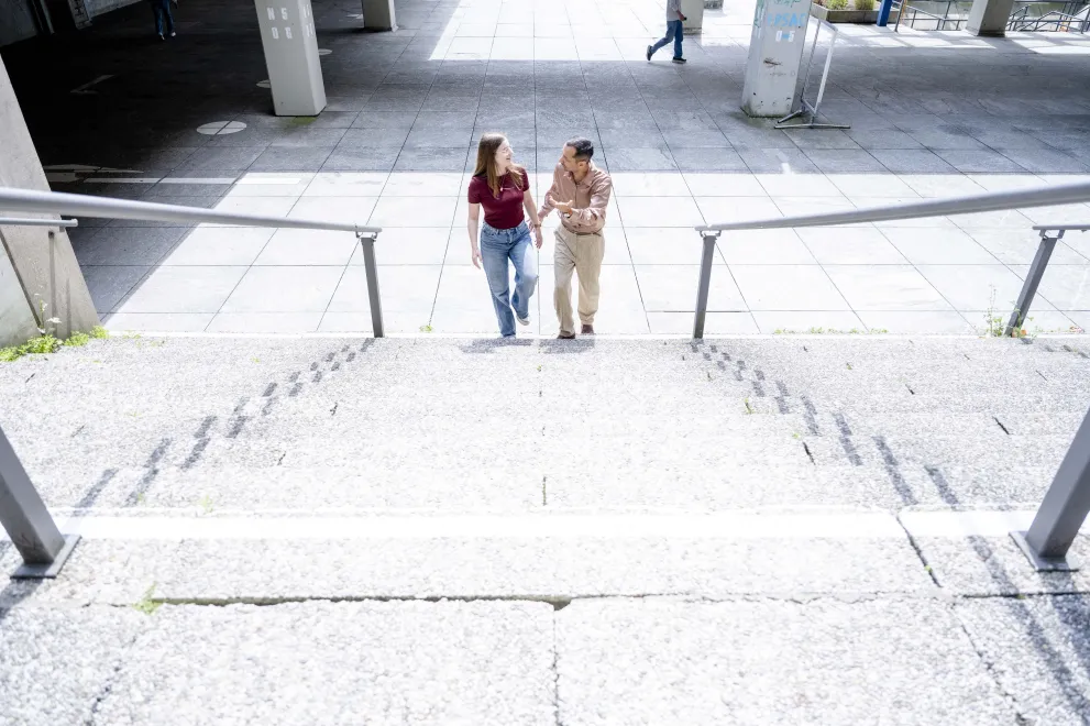 Foto: ZWei Personen, eine männliche und eine weiblich gelesene, steigen ein Treppe hoch und unterhalten sich dabei. Das Bild ist von dem oberen Teil der Treppe aufgenommen.
