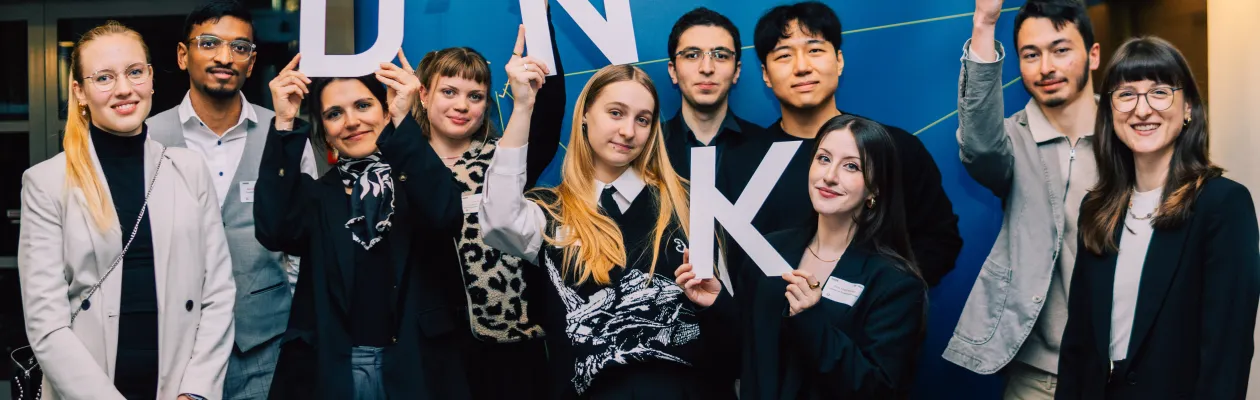 Foto: Mehrere Personen stehen vor einem blauen Aufsteller. Auf dem Aufsteller sind das Logo des Deutschlandstipendiums und das Logo der Ruhr-Universität Bochum zu sehen. Die Personen halten die Buchstaben D, A, N, K und E hoch. Das ergibt das Wort "Danke".