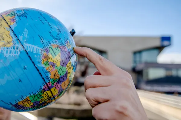 Foto: Groß im Vordergrund eine Hand, die mit einem Finger auf einen Globus zeigt; im Hintergrund der RUB-Campus, erkennbat am verschwommen Logo am Musischen Zentrum.