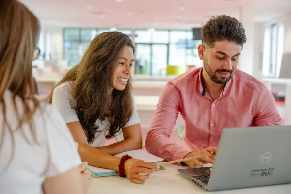 Foto: Drei Studierende sitzen an einem Tisch. Eine Person zeigt mit einem Stift auf einen Laptop.