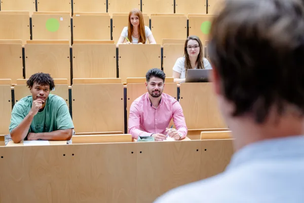 Foto: Ein Dozent steht in einem Hörsaal vor einer Lerngruppe.