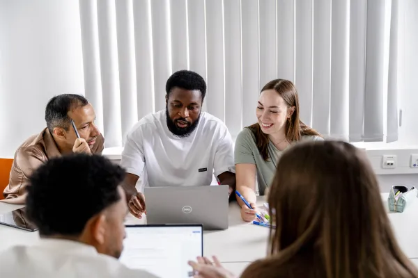 Foto: Mehrere Personen sitzen an einem Tisch mit Laptops und Stiften. Sie unterhalten sich, schauen auf die Laptops und scheinen zu überlegen.