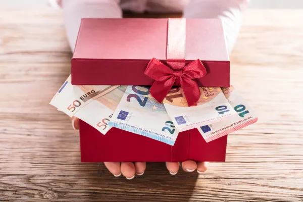 Foto: Zwei Hände halten eine rote Geschenkbox mit roter Schleife. Der Deckel der Box ist leicht geöffnet. Durch den Deckel sieht man Geldscheine.