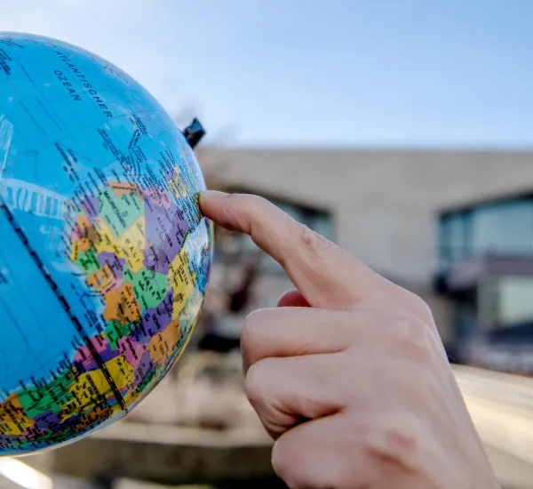 Foto: eine HAnd zeigt auf einen Globus, im Hintergrund der RUB-Campus mit Logo an einem Gebäude